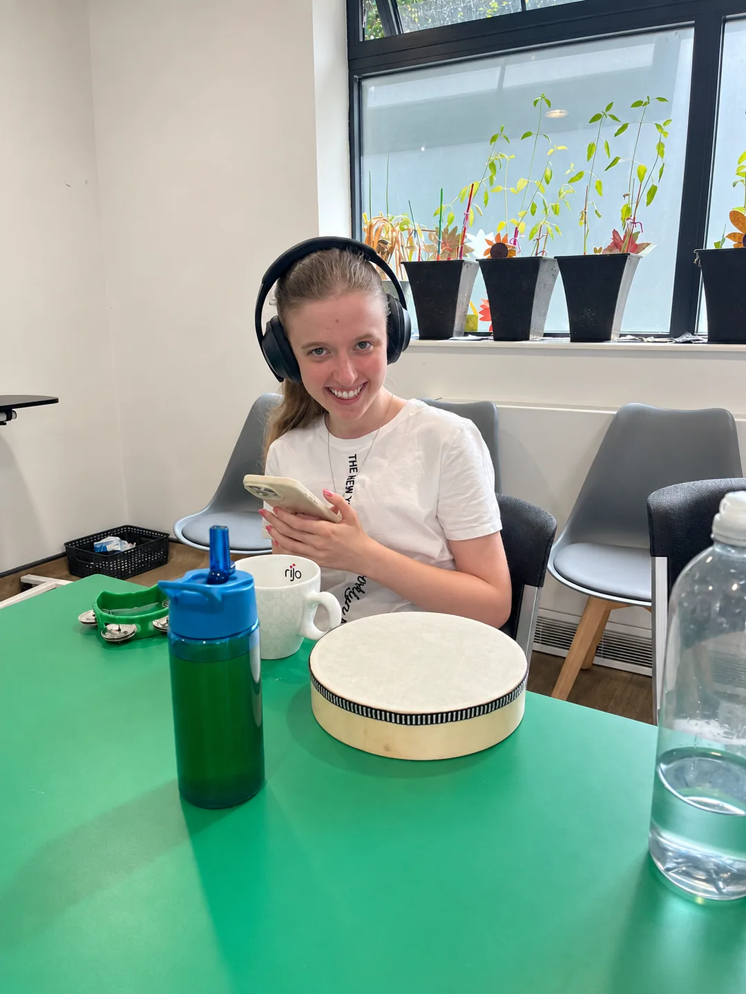 A young woman in headphones smiling over her phone, a hand drum and tambourine on the green table in front of her
