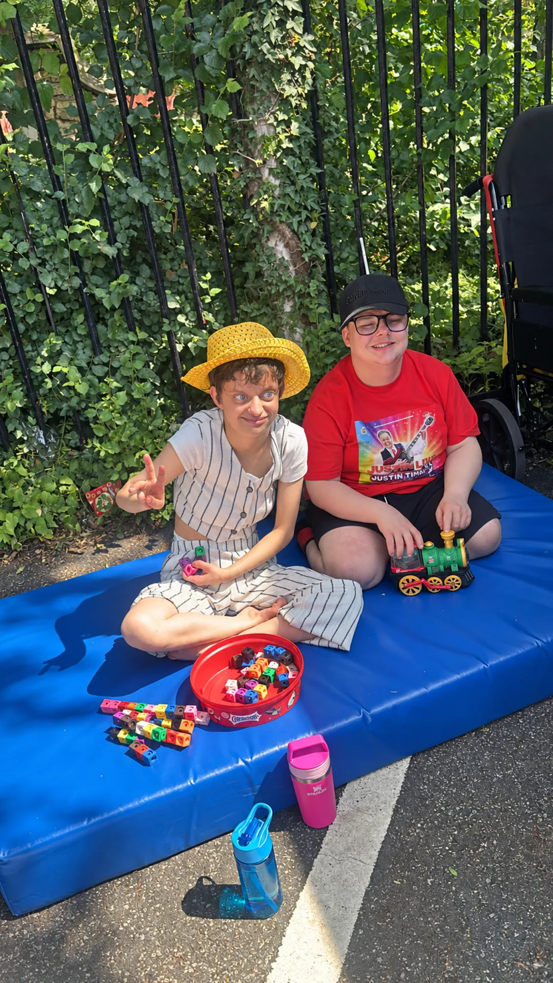 Two members sitting on a blue mat outside, one in a yellow straw hat making a peace sign