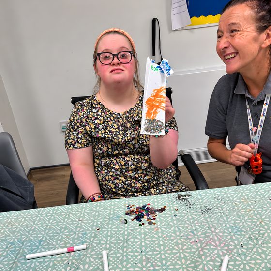 A member holding up a paper creation she decorated, sitting beside a smiling staff member at the craft table.